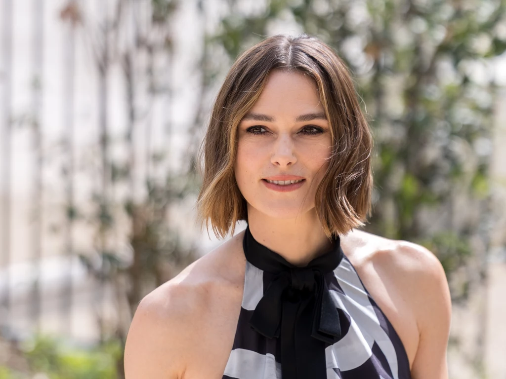 Aktorka Keira Knightley w modnym bobie Kobieta z krótkimi, brązowymi włosami w czarno-białej sukience na tle rozmytego ogrodu lub parku, patrząca wprost z delikatnym uśmiechem.
