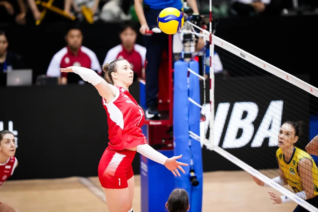 Liga Narodów siatkarek. Aleksandra Gryka rozpoczęła mecz Polska - Brazylia w podstawowym składzie Siatkarka w czerwonym stroju wyskakuje przy siatce, przygotowując się do ataku, naprzeciw zawodniczka w żółtym stroju próbująca blokować piłkę. W tle widoczni sędziowie oraz osoby obserwujące mecz.