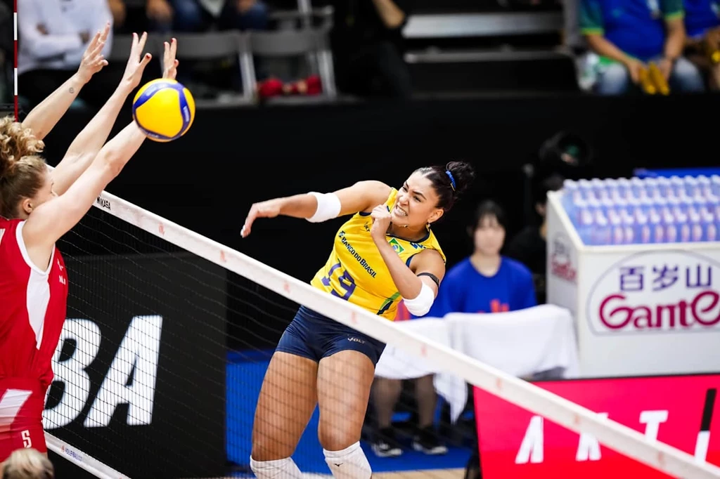 Liga Narodów siatkarek. Tainara w trakcie meczu Polska - Brazylia Zawodniczka siatkarska drużyny w żółtym stroju dynamicznie atakuje piłkę ponad siatką, próbując zdobyć punkt, podczas gdy zawodniczka drużyny przeciwnej w czerwonym stroju blokuje atak, a w tle widoczni są sędziowie i ławka rezerwowych.