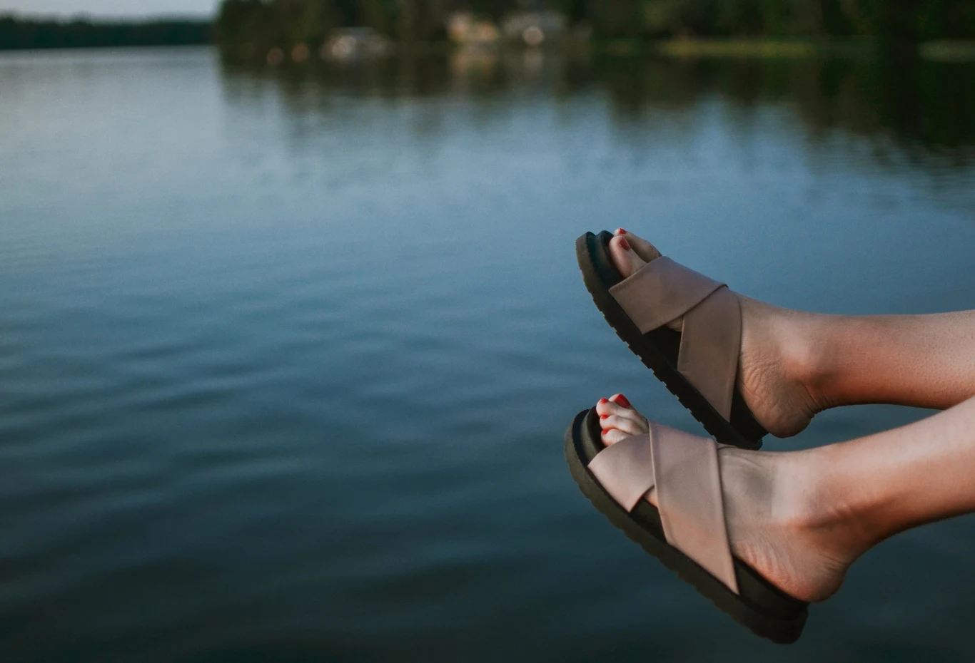 Czy w tym roku możemy zdaniem jasnowidza liczyć na ładne lato? Stopy w sandałach wystawione nad wodą jeziora, w tle widoczny spokojny brzeg i zabudowania na drugim planie.