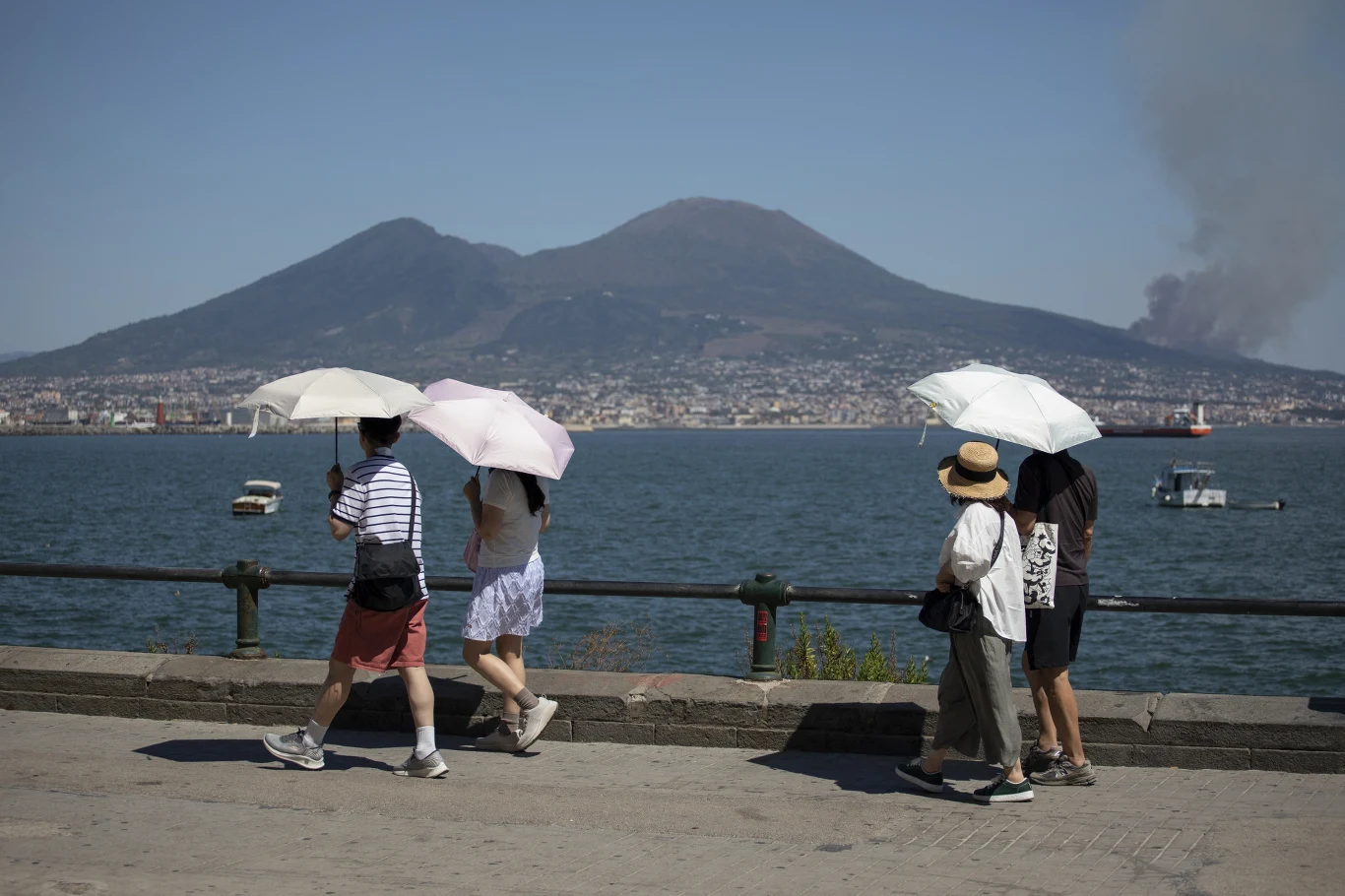 Turyści w Neapolu patrzą na pożar w oddali Cztery osoby spacerują nad brzegiem morza, trzymając białe i jasne parasole chroniące przed słońcem, w tle widoczny masyw wulkanu oraz dym unoszący się po prawej stronie, a na wodzie pływają łodzie.