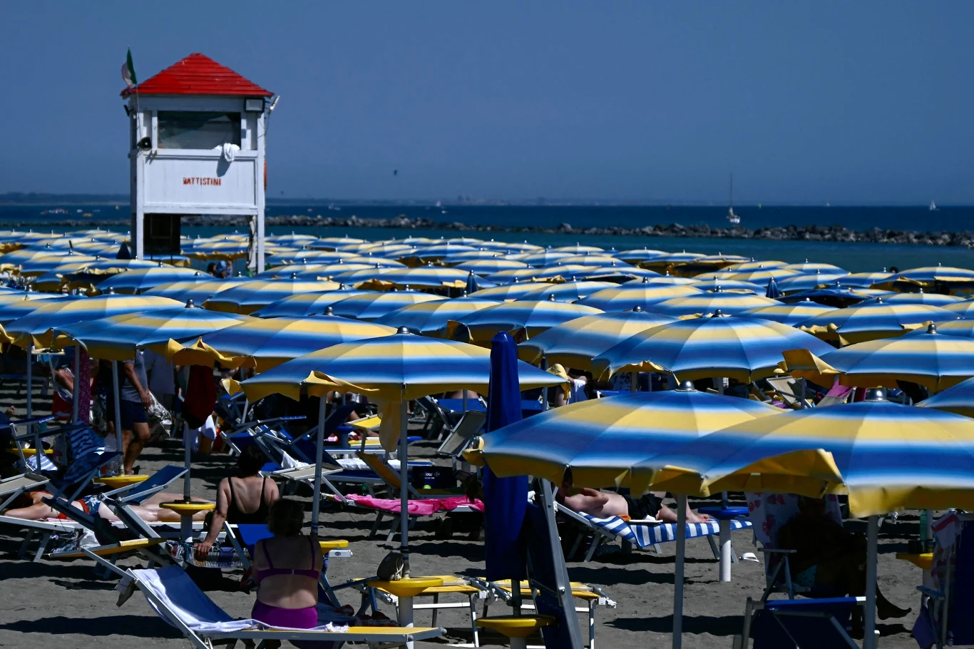 Plaża w Fiumicino we Włoszech Tłum ludzi relaksujących się pod niebiesko-żółtymi parasolami plażowymi, w tle wieża ratownika z czerwonym dachem oraz spokojne morze.