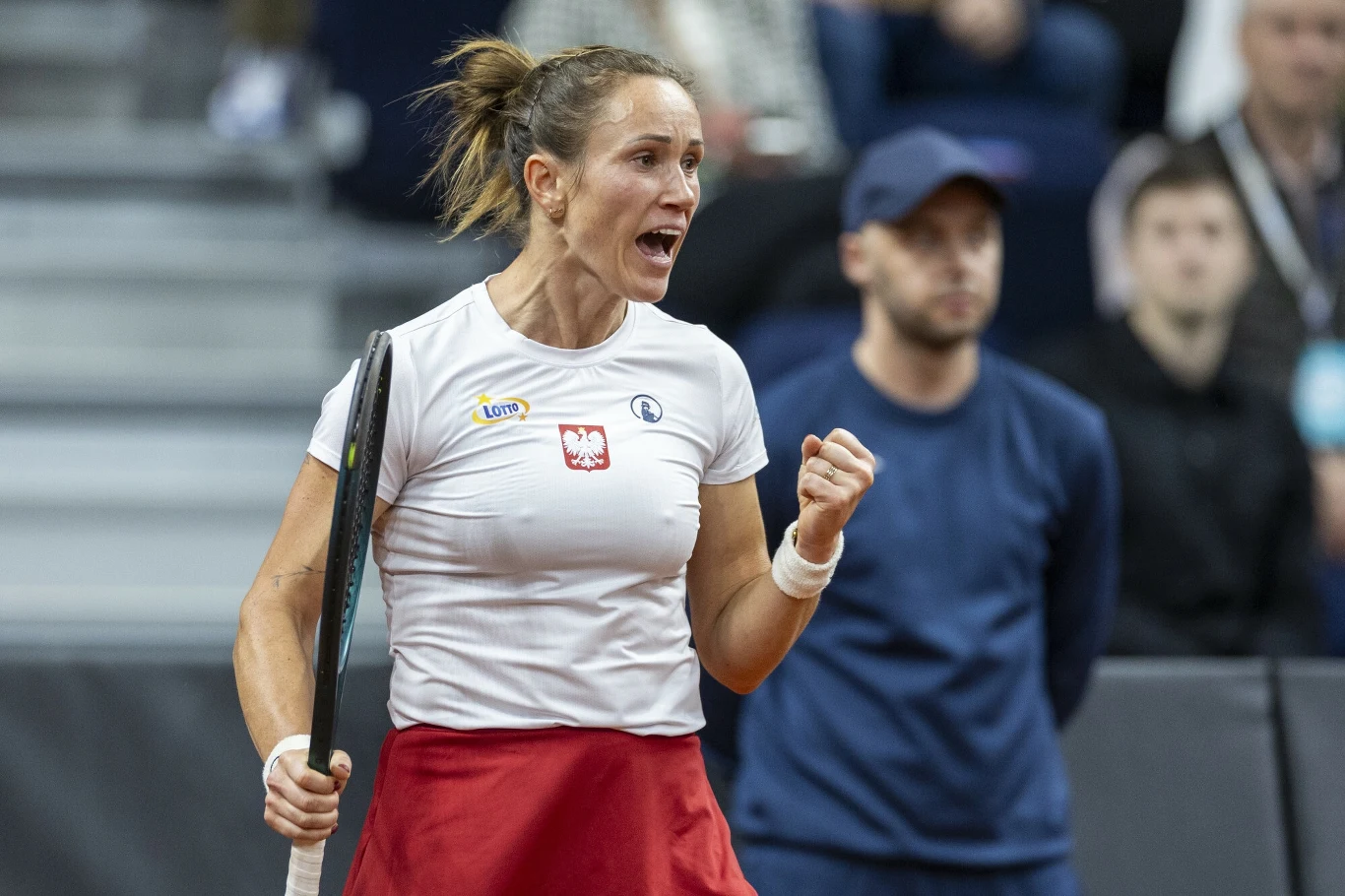 Katarzyna Kawa Zawodniczka tenisa w trakcie meczu, ubrana w biało-czerwoną koszulkę z polską flagą, wyraża mocne emocje, zaciskając pięść i krzycząc z triumfem, w tle widzowie oraz osoba w niebieskiej bluzie i czapce.