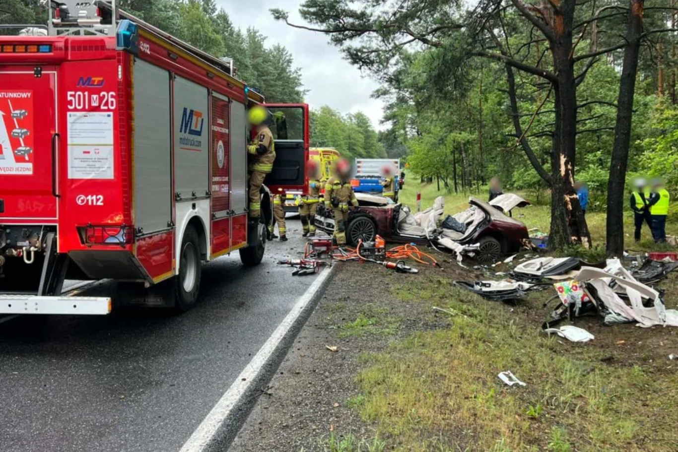 Strażacy odcięli dach, aby wydostać z wnętrza pojazdu 74-letnią kobietę. Strażacy i służby ratunkowe pracują przy wraku samochodu rozbitego o drzewo, na poboczu rozrzucone są części pojazdu, w tle na leśnej drodze stoją kolejne pojazdy służbowe oraz kilka osób.