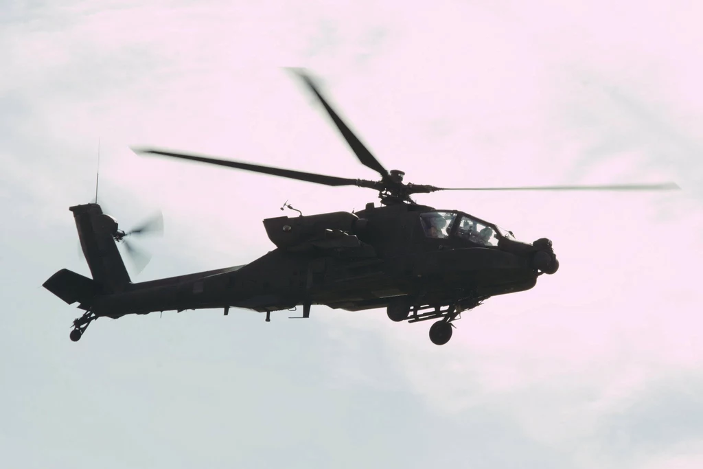 AH-64D w 56. Bazie Lotniczej w Latkowie Ciemny helikopter wojskowy z wirującymi wirnikami lecący na tle jasnego, lekko zachmurzonego nieba, widoczny głównie z profilu
