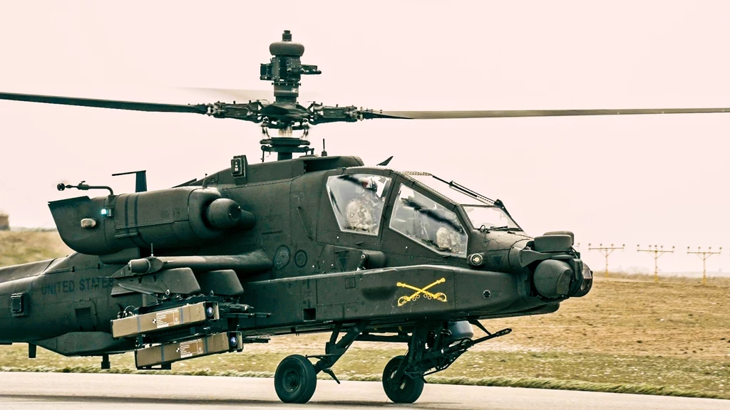 Amerykański AH-64 Apache z podwieszonymi pociskami Spike NLOS Śmigłowiec bojowy AH-64 Apache z widocznym uzbrojeniem pod skrzydłami, oznaczeniami wojsk lądowych Stanów Zjednoczonych, stojący na płycie lotniska na tle pustego terenu.
