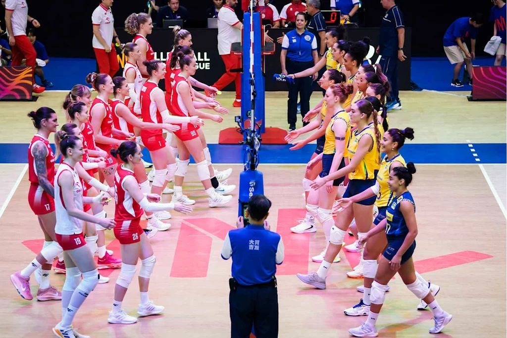 Liga Narodów siatkarek. Polska przegrała z Brazylią Dwie żeńskie drużyny siatkarskie w strojach sportowych witają się przed rozpoczęciem meczu, po przeciwnych stronach siatki, sędzia stoi z tyłu na środku boiska, na trybunach widoczna obsługa techniczna i kilku widzów.