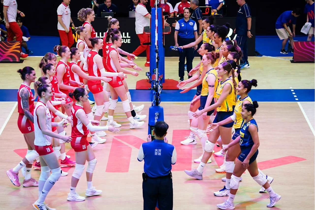 Dwie żeńskie drużyny siatkarskie w strojach sportowych witają się przed rozpoczęciem meczu, po przeciwnych stronach siatki, sędzia stoi z tyłu na środku boiska, na trybunach widoczna obsługa techniczna i kilku widzów.
