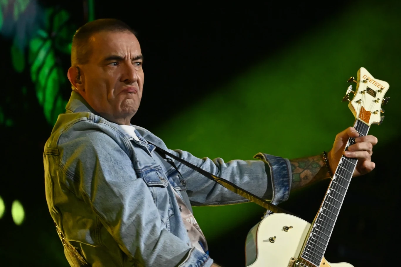 Andrzej Kraiński z zespołu Kobranocka Mężczyzna w dżinsowej kurtce gra na białej gitarze elektrycznej, wykonując energiczny ruch podczas występu na scenie; w tle zielone światła sceniczne podkreślają koncertową atmosferę.