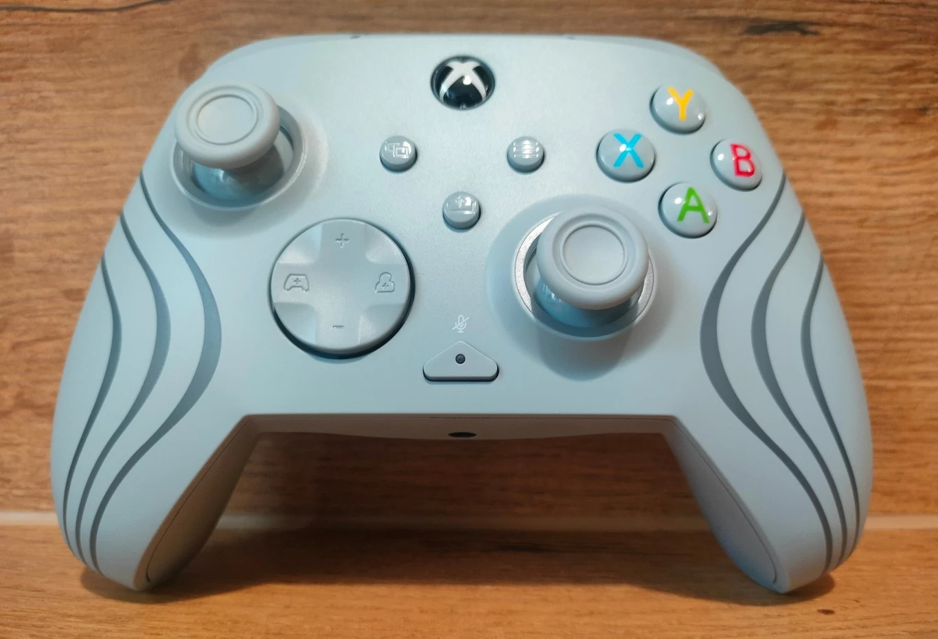 Turtle Beach Afterglow Wave Szary kontroler do gier z kolorowymi przyciskami i dwoma drążkami analogowymi, umieszczony na drewnianej powierzchni.