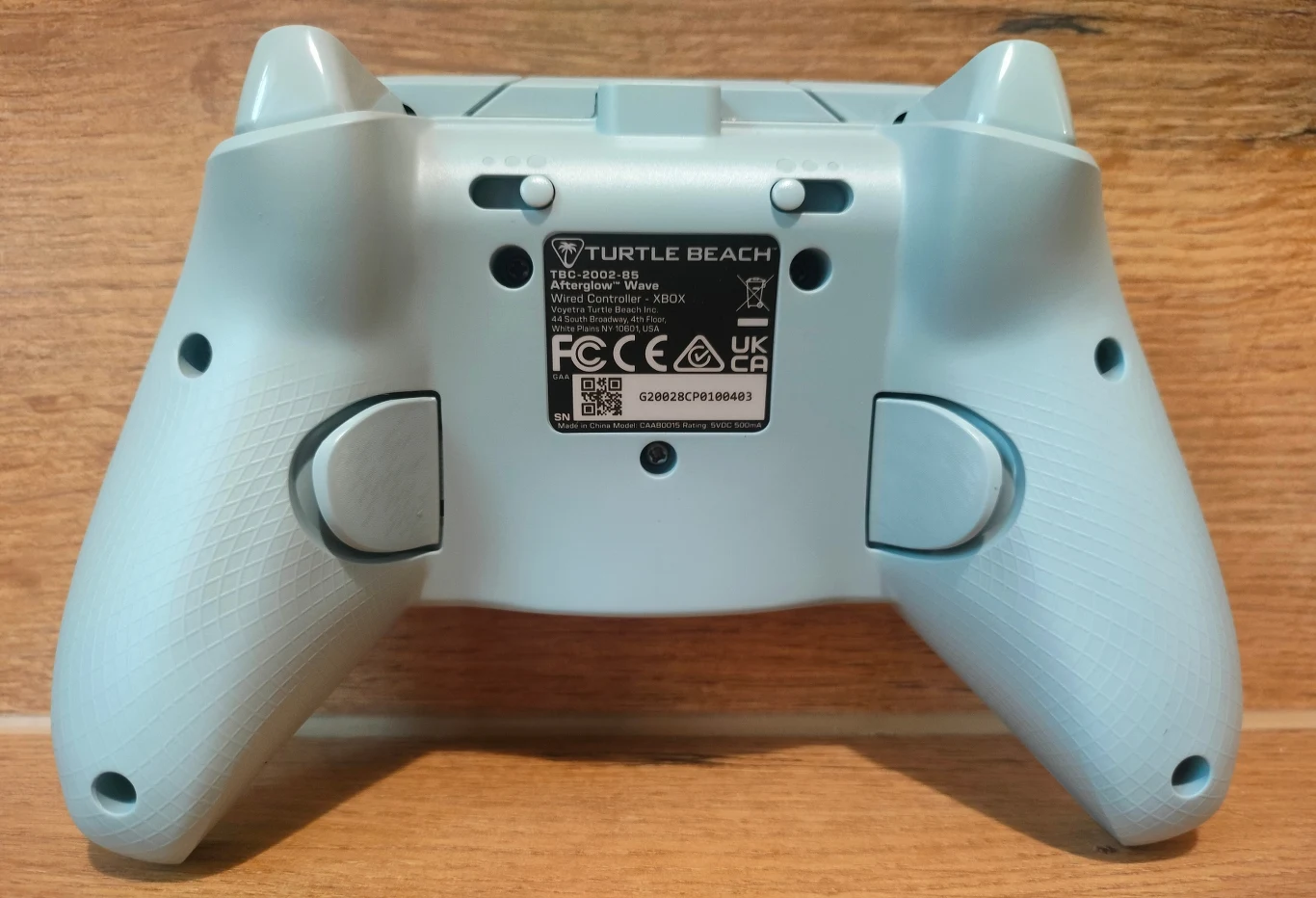 Turtle Beach Afterglow Wave Kontroler do gier Turtle Beach w jasnym kolorze, widok od tyłu z wyraźnie widoczną tabliczką znamionową i przyciskami na uchwytach, na drewnianym tle.