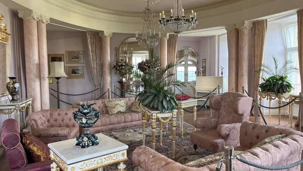 Elegancki salon urządzony w stylu klasycznym z różowymi, welurowymi kanapami, złotymi detalami, kryształowymi żyrandolami, ozdobnymi roślinami oraz dużym lustrem odbijającym światło dzienne wpadające przez wysokie okna.