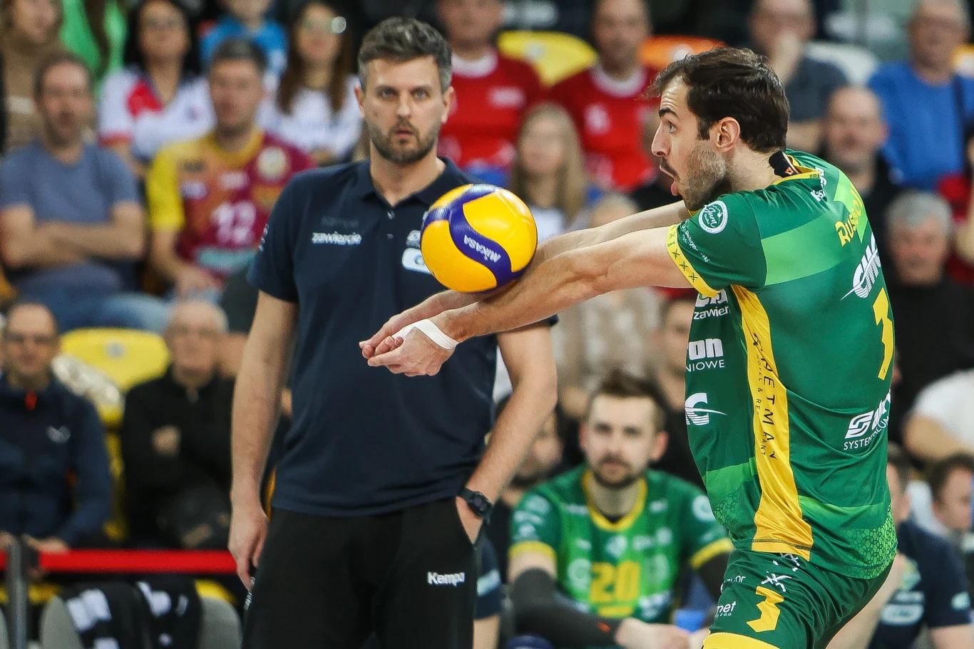 Aaron Russell i Michał Winiarski (w tle) Zawodnik siatkarski w zielono-żółtym stroju odbijający piłkę, skupiony trener w tle oraz kibice na trybunach.