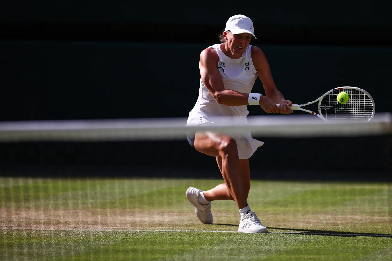 Tenisistka w białym stroju wykonuje odbicie piłki podczas meczu na trawiastym korcie w pełnym słońcu.