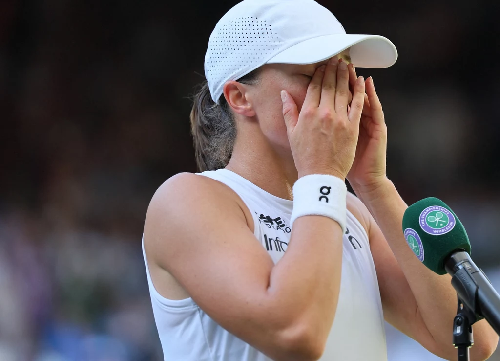 Iga Świątek, Wimbledon 2025 Tenisistka ubrana w biały strój i czapkę z daszkiem stoi przy mikrofonie, zakrywając twarz dłońmi, sprawiając wrażenie wzruszonej lub przeżywającej silne emocje, w tle widoczne zamazane sylwetki i elementy kortu tenisowego.