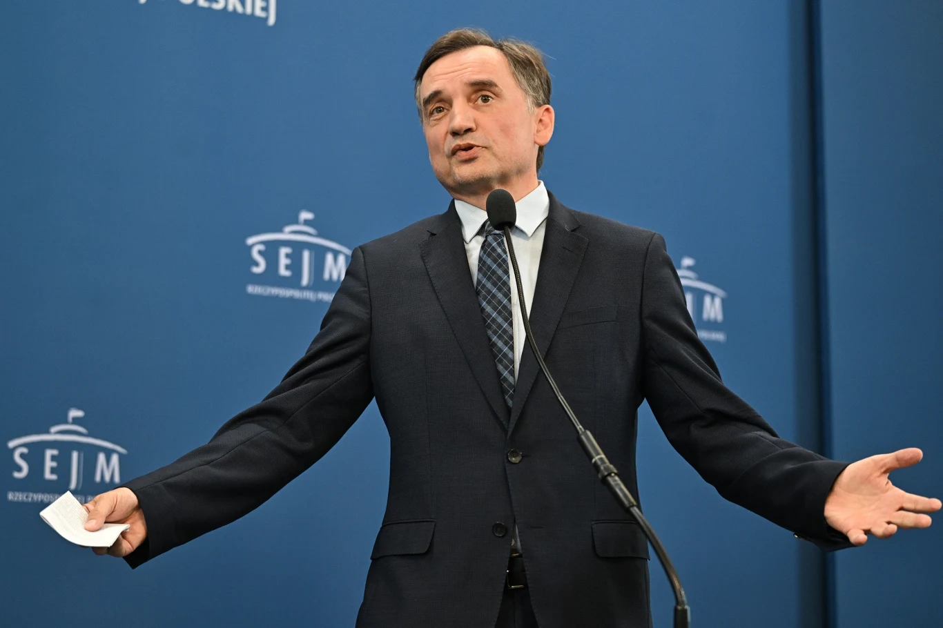 Zbigniew Ziobro Mężczyzna w garniturze stoi przy mikrofonie podczas konferencji prasowej na tle niebieskiej ścianki z napisem Sejm Rzeczypospolitej Polskiej, trzyma w ręku kartkę papieru, wykonuje gest otwartymi dłońmi.