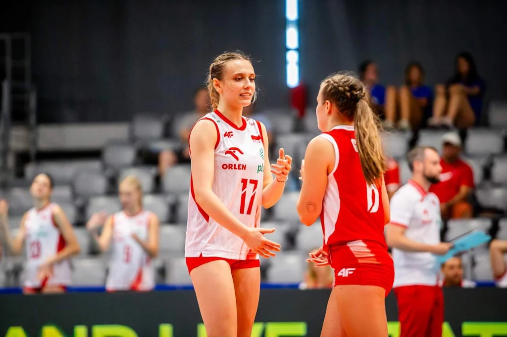 Dwie zawodniczki siatkarskie drużyny Polski w sportowych strojach rozmawiają na boisku podczas meczu, w tle widoczni są inni członkowie drużyny oraz kibice na trybunach.