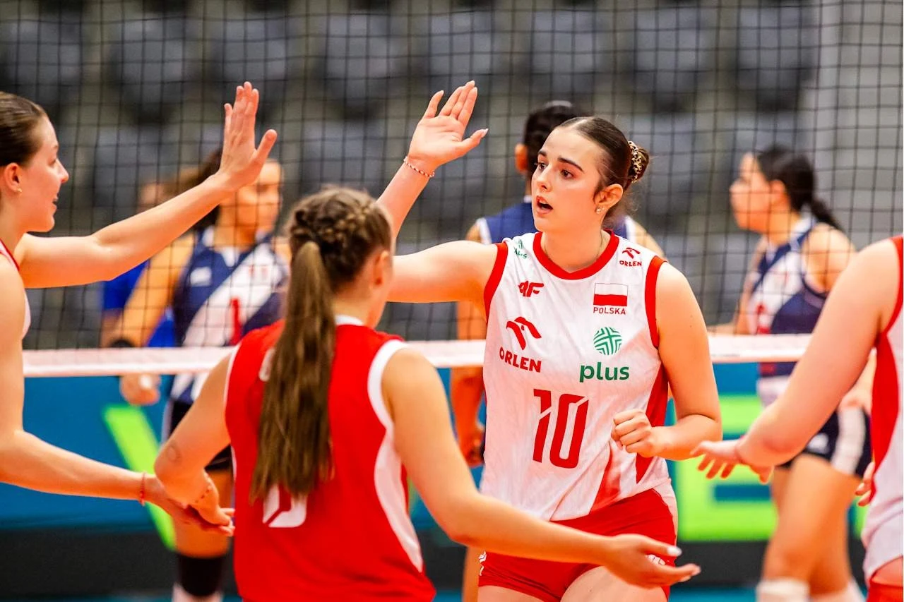 Siatkarki reprezentacji Polski do lat 19 w czasie mistrzostw świata Siatkarki w biało-czerwonych strojach świętują udaną akcję na boisku podczas meczu; w tle widoczne siatka oraz zawodniczki drużyny przeciwnej.