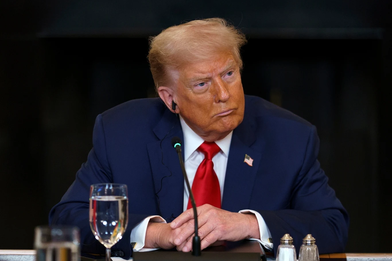 Mężczyzna w średnim wieku w granatowym garniturze i czerwonym krawacie siedzi przy stole z mikrofonem, przed nim stoi szklanka wody i przyprawniki, w tle widoczne ciemne wnętrze.