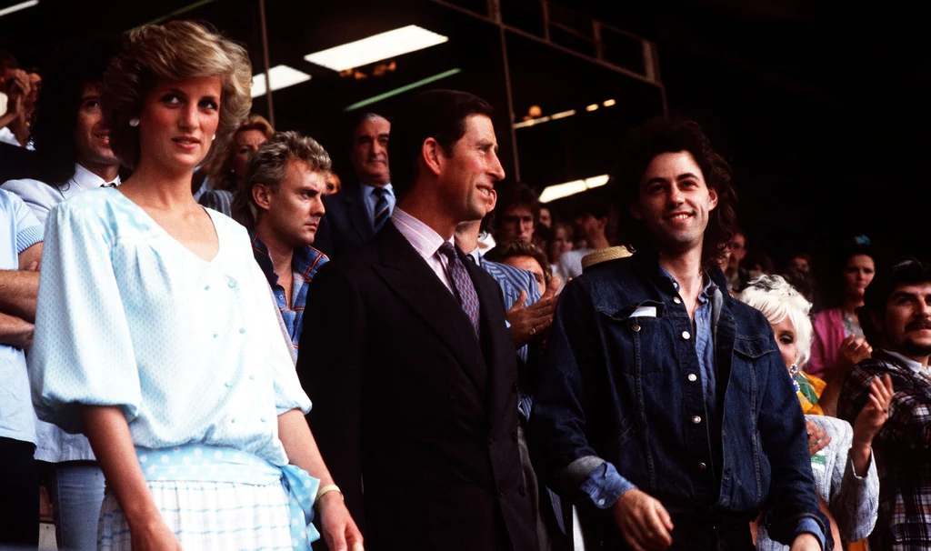 Na Wembley pojawili się ówczesny książę Karol (obecny król) z żoną, księżną Dianą. Z prawej jeden z organizatorów Live Aid - Bob Geldof Grupa osób dobrze ubrana, w centrum elegancka kobieta w jasnej sukience, obok niej mężczyzna w garniturze i młody mężczyzna w dżinsowej kurtce. W tle inne osoby stojące i uczestniczące w wydarzeniu publicznym, atmosfera oficjalna i pozytywna.