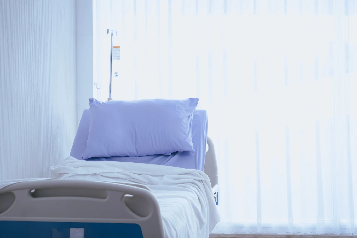 Pacjent zakażony dżumą zmarł w szpitalu w Arizonie Łóżko szpitalne ustawione przy oknie zasłoniętym białą firanką, na łóżku ułożona biała pościel i niebieska poduszka, obok widoczny stojak na kroplówkę.