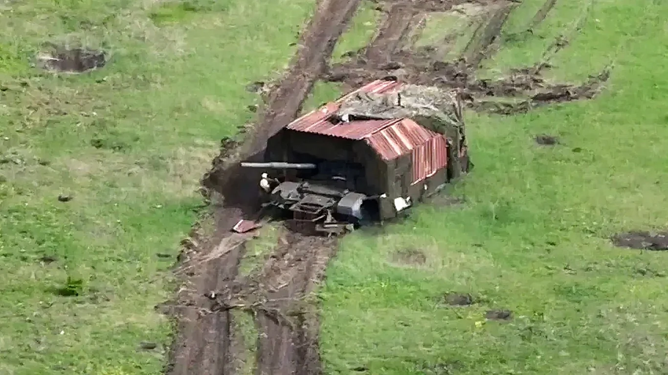 Stalowe "czołgi-żółwie" pojawiły się na froncie w Ukrainie Czołg częściowo ukryty pod pokryciem z elementów blachy falistej stoi na zielonym polu, wokół widoczne są ślady gąsienic na ziemi i rozjeżdżona trawa.