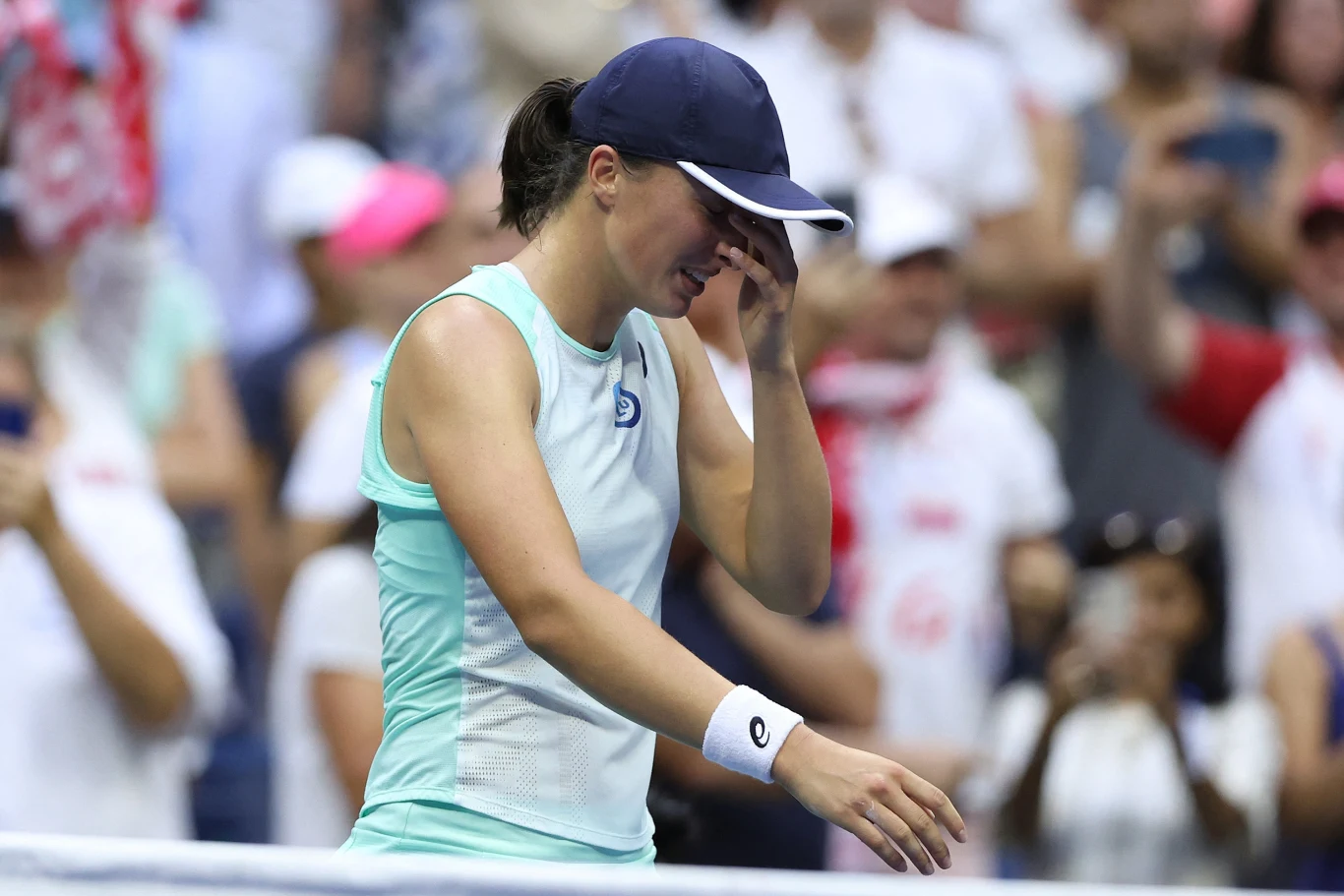 Tenisistka w sportowym stroju obejmuje ręką twarz i opuszcza głowę, stojąc na korcie tenisowym, w tle tłum kibiców.