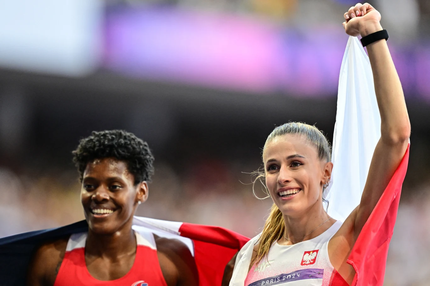 Dwie uśmiechnięte sportsmenki biegnące z flagami swoich krajów na stadionie lekkoatletycznym, kobieta po prawej trzyma biało-czerwoną flagę z godłem, a druga zawodniczka okryta jest flagą w czerwono-biało-niebieskich barwach.