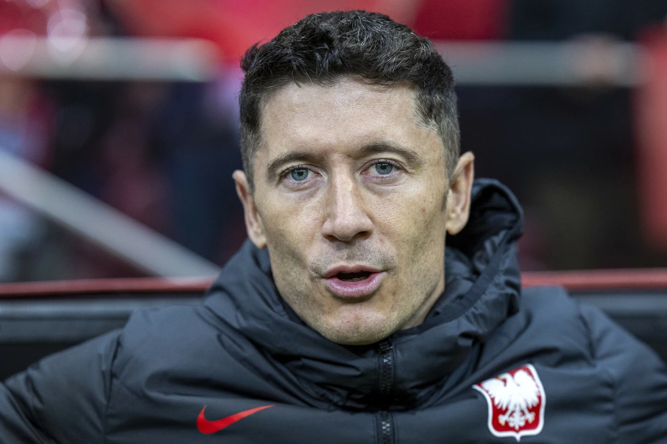 Zbliżenie na mężczyznę w czarnej kurtce z emblematem polskiego orła na piersi, mającego krótkie ciemne włosy i poważny wyraz twarzy. Tło rozmyte, dominują czerwone barwy.