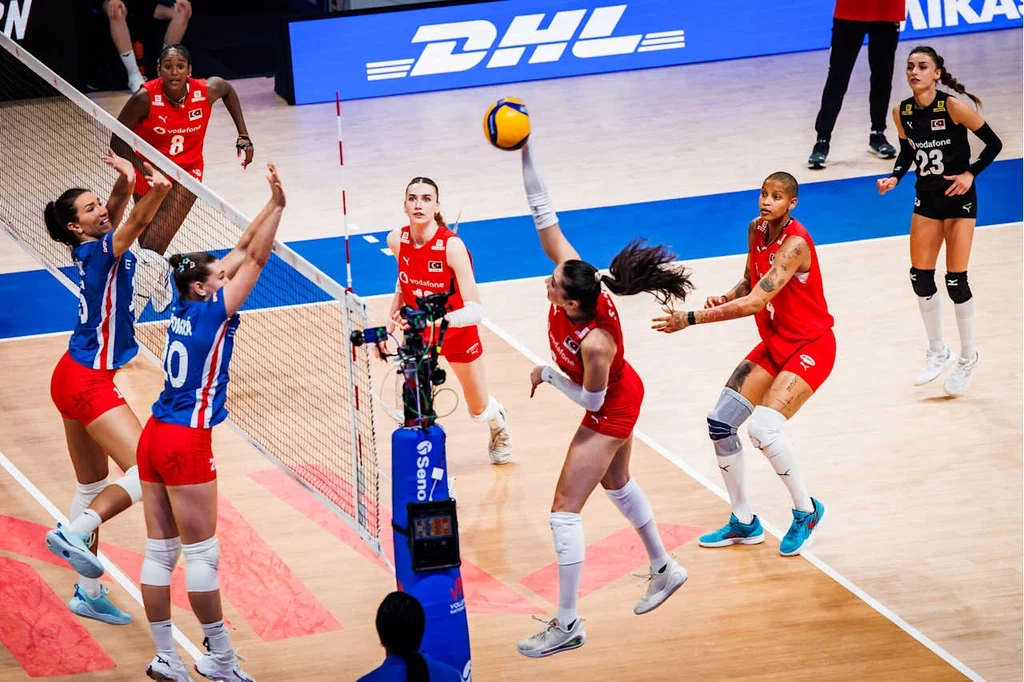 Mecz Czechy - Turcja w Lidze Narodów Zawodniczki dwóch drużyn siatkarskich rywalizują przy siatce, jedna z nich próbuje przebić piłkę na stronę przeciwniczek, inne zawodniczki przygotowują się do akcji, a w tle widoczny jest sędzia oraz reklama DHL.