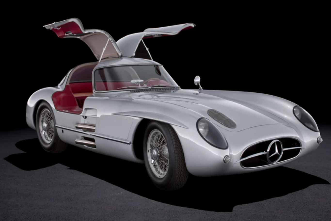 Srebrny, klasyczny samochód sportowy z otwartymi drzwiami typu gullwing i czerwonym wnętrzem, na czarnym tle.