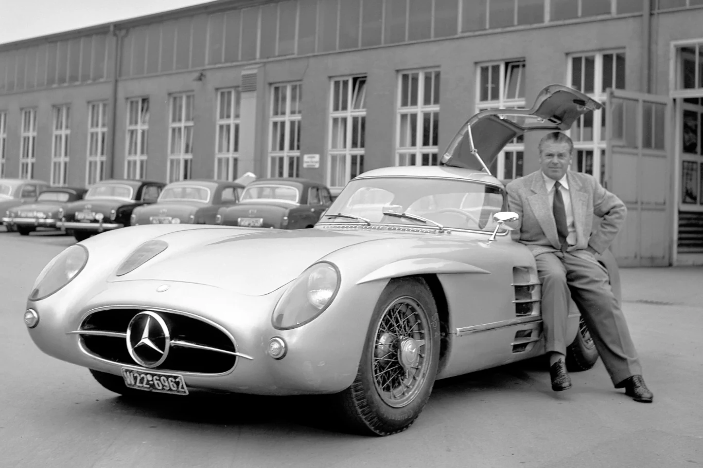 Mężczyzna w garniturze opiera się o zabytkowy samochód sportowy marki Mercedes-Benz z unoszonymi do góry drzwiami typu gullwing, na tle budynku fabrycznego oraz zaparkowanych klasycznych aut.