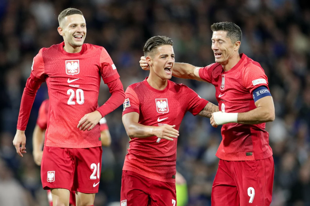 Sebastian Szymański, Kacper Urbański, Robert Lewandowski Trzech piłkarzy polskiej reprezentacji w czerwonych strojach świętuje na murawie stadionu, uśmiechnięci z widoczną radością po strzeleniu bramki lub wygranej akcji.