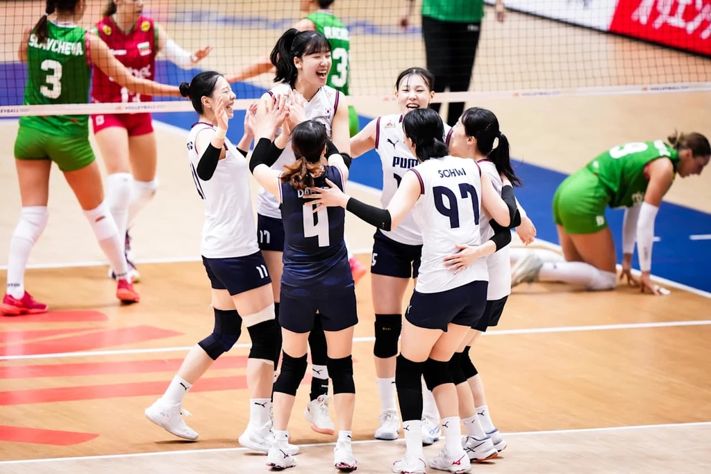Siatkarki reprezentacji Korei Południowej Zawodniczki drużyny siatkarskiej świętujące zdobycie punktu, ubrane w stroje meczowe, na tle hali sportowej z widoczną linią siatki oraz drużyną przeciwną w zielonych strojach.