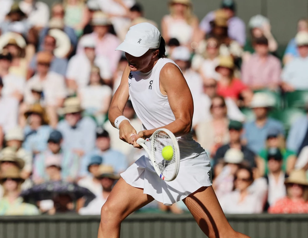 Iga Świątek, Wimbledon 2025 Tenisistka w białym stroju odbijająca piłkę podczas meczu na korcie trawiastym, publiczność w tle śledzi rozegranie akcji.