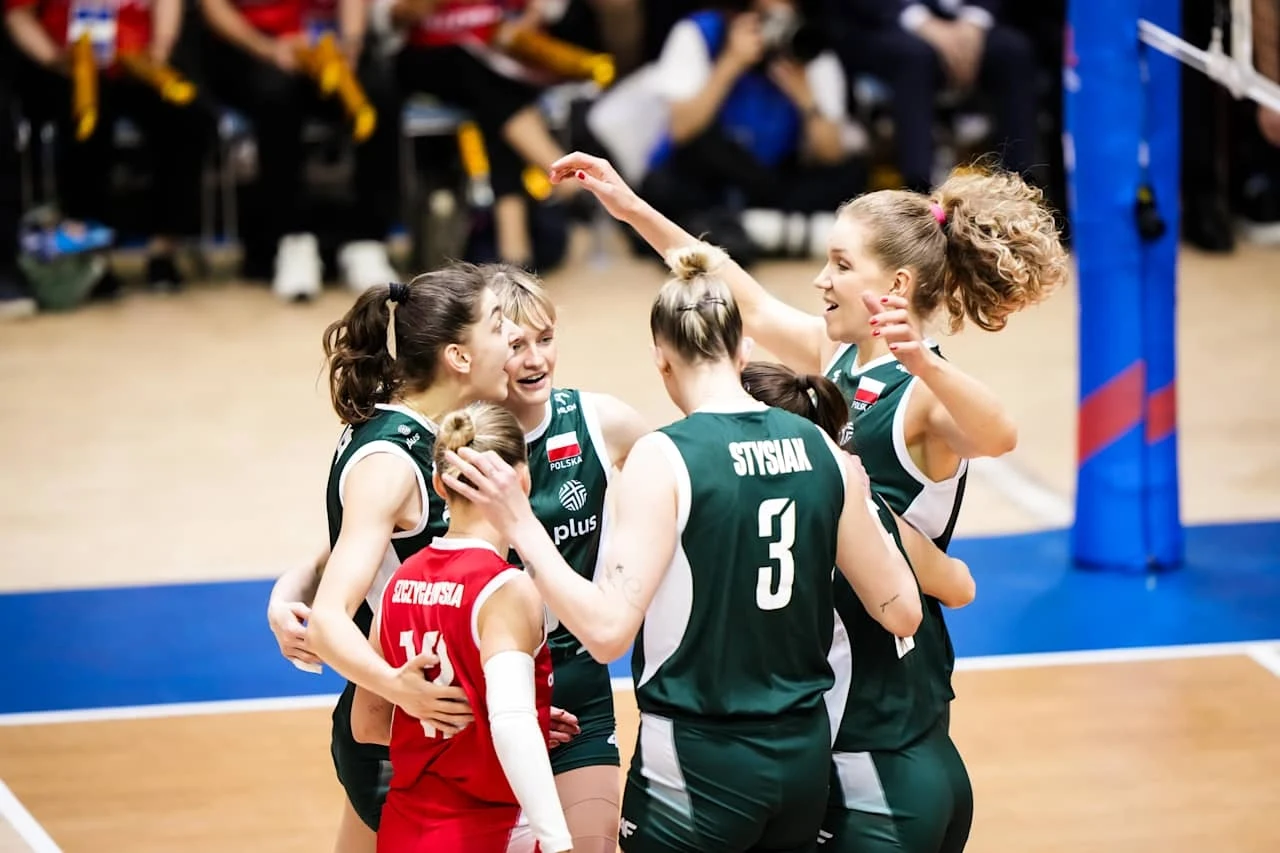 Grupa siatkarek w zielonych i czerwonych strojach świętuje zdobycie punktu podczas meczu, przybijając sobie piątki i uśmiechając się nawzajem, w tle widoczni kibice oraz fragment siatki.
