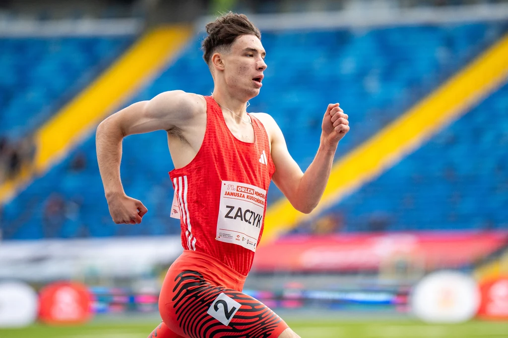 Łukasz Zaczyk Lekkoatleta w czerwonym stroju biegnący po bieżni stadionu podczas zawodów, w tle widoczne puste niebieskie trybuny.