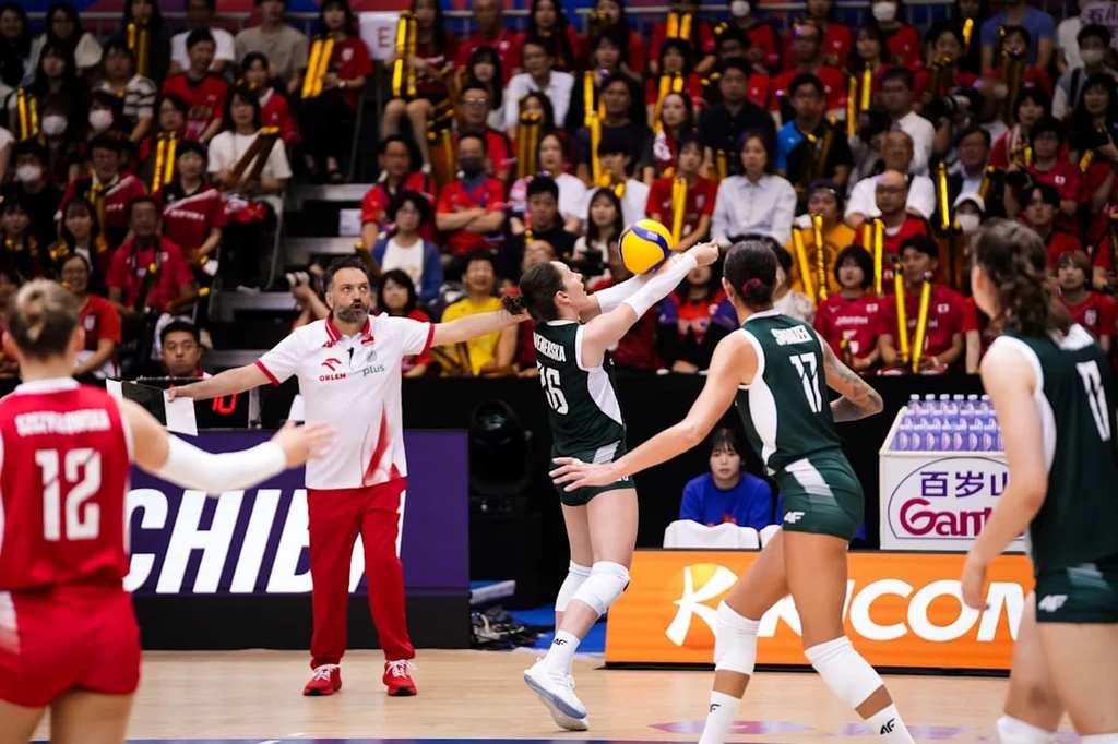 Liga Narodów siatkarek. Kadr z meczu Polska - Japonia Zawodniczki dwóch drużyn siatkarskich rywalizują na boisku podczas dynamicznej akcji, w tle trener wydaje polecenia, a trybuny są wypełnione kibicami.