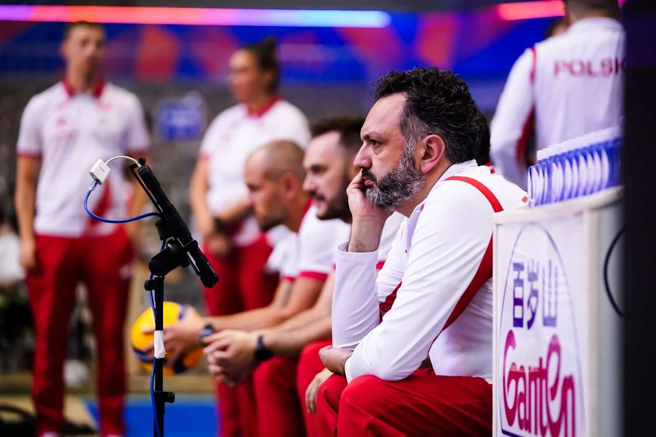 Stefano Lavarini, trener polskich siatkarek Trener i sztab szkoleniowy reprezentacji Polski w siatkówce siedzący na ławce rezerwowych podczas meczu, ubrani w białe koszulki z czerwonymi elementami, z widocznym napięciem na twarzach niektórych osób.