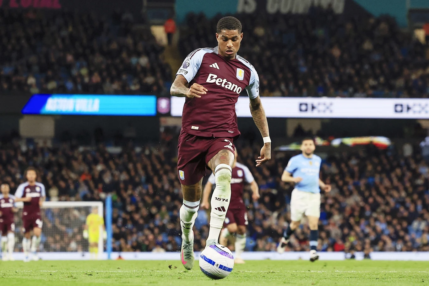 Zawodnik drużyny Aston Villa w bordowo-niebieskim stroju prowadzi piłkę podczas meczu, w tle widać innych piłkarzy oraz publiczność na stadionie.