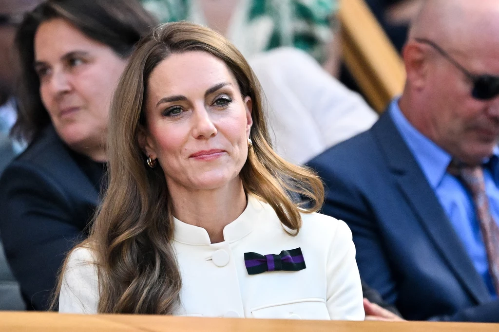 Księżna Kate na finale singla kobiet, Wimbledon 2025 Kobieta w eleganckiej kremowej marynarce z przypiętą broszką w kształcie kokardy siedzi na trybunach, w tle inne osoby obserwujące wydarzenie.
