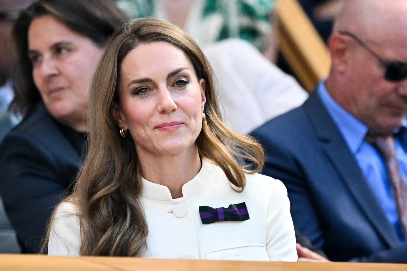 Księżna Kate na finale singla kobiet, Wimbledon 2025 Kobieta w eleganckiej kremowej marynarce z przypiętą broszką w kształcie kokardy siedzi na trybunach, w tle inne osoby obserwujące wydarzenie.
