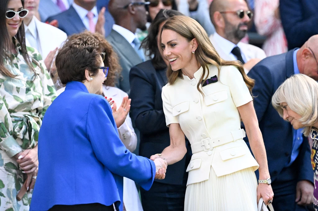 Księżna Kate i Billie Jean King na finale singla kobiet, Wimbledon 2025 Dwie kobiety ściskające sobie dłonie w otoczeniu ludzi ubranych elegancko, jedna z nich ma białą sukienkę, a druga niebieską marynarkę, w tle widać liczną publiczność.
