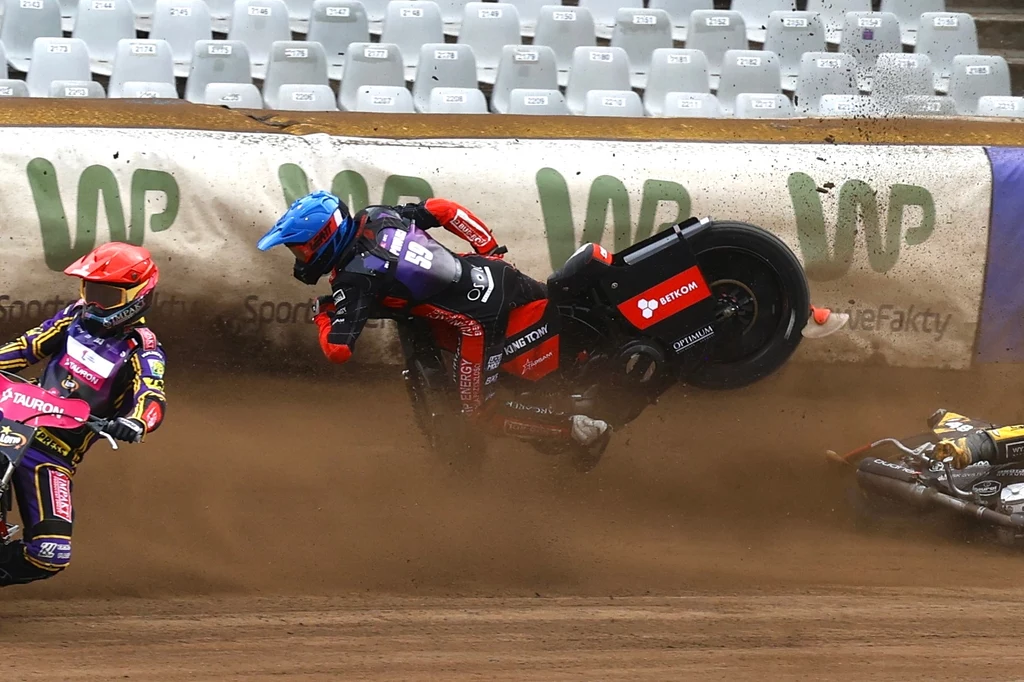 Moment upadku Przemysława Pawlickiego w SEC w Bydgoszczy Motocyklista w niebieskim kasku wypada z toru wraz z motocyklem podczas wyścigu żużlowego, na torze widoczni także inni zawodnicy oraz trybuny z pustymi miejscami.