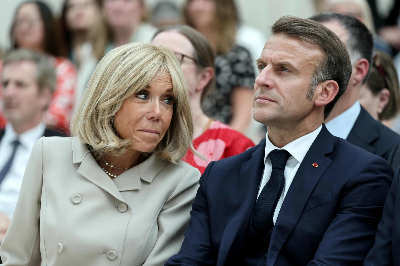 Brigitte i Emmanuel Macron podczas wizyty w Wielkiej Brytanii Para w eleganckich ubraniach siedzi obok siebie wśród tłumu, mężczyzna w garniturze i krawacie patrzy poważnie w bok, kobieta w jasnym płaszczu skierowana w jego stronę, w tle rozmyte postacie innych osób.