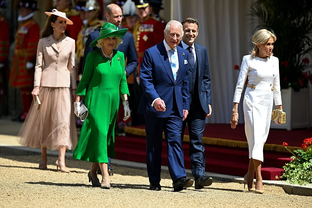 Brigitte i Emmanuel Macron , król Karol III i królowa Kamila, książę William i księżna Kate Kilka elegancko ubranych osób idzie wzdłuż ogrodowego chodnika, mężczyzna w niebieskim garniturze znajduje się na środku grupy, towarzyszą mu kobiety w wyrafinowanych strojach oraz inni mężczyźni, w tle widać strażników w ceremonialnych mundurach i cze...