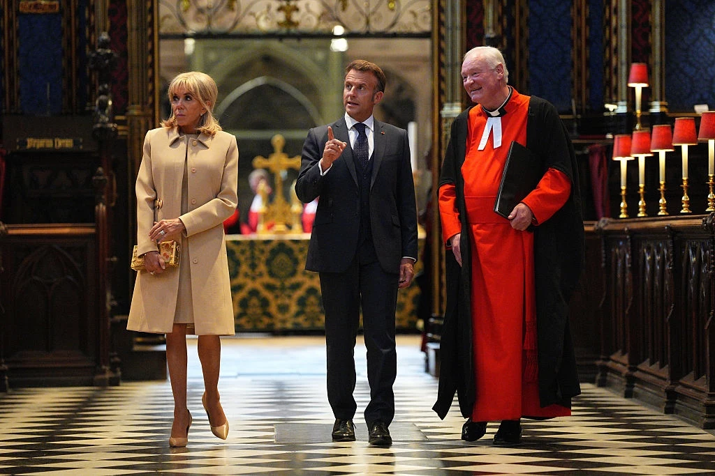 Emmanuel i Brigitte Macron w opactwie westminsterskim Trzy osoby w eleganckich strojach spacerują po wnętrzu bogato zdobionej, historycznej budowli z charakterystyczną, czarno-białą posadzką i licznymi świecami po prawej stronie.