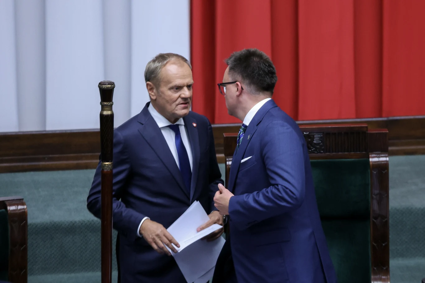 Relacje między Donaldem Tuskiem i Szymonem Hołownią są ostatnio wyjątkowo napięte Dwóch mężczyzn w garniturach prowadzi rozmowę w sali sejmowej, po lewej widoczny fragment mównicy, w tle dekoracja z pionowych pasów kolorów biały, szary i czerwony.