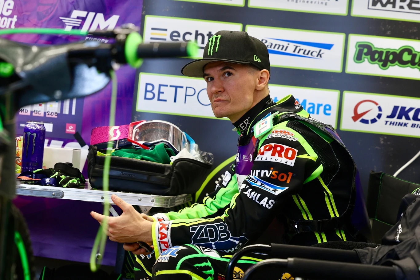 Żużlowiec w profesjonalnym kombinezonie sportowym siedzący na ławce w strefie serwisowej, otoczony sprzętem i banerami sponsorów.