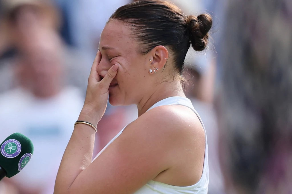 Amanda Anisimova Kobieta w sportowym stroju z zamkniętymi oczami zasłania twarz dłonią, wyrażając silne emocje. W tle rozmyty tłum i mikrofon z logo turnieju tenisowego sugerują, że znajduje się na korcie podczas ważnego wydarzenia.